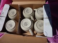 Top view of six stoneware mugs placed upside down in a cardboard box, showing the manufacturer markings on the base.