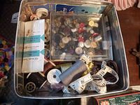 Photo showing tin box with assorted sewing items including buttons in plastic container, thread spools, measuring tape, and packaged trims.