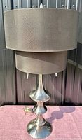 Full view of the Mid-Century Modern lamp showing the brushed stainless steel base and the double drum fabric shade in beige/taupe color.