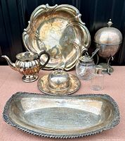 Photo showing a set of seven old silver plated items arranged on a surface, including large floral shaped tray, teapot, egg coddler, platter, glass jar and containers.