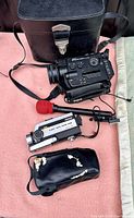 Black Yashica movie camera with control dials and attached microphone with red foam wind muff, next to a black hard carrying case with shoulder strap.