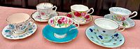 Image showing six vintage porcelain cup and saucer sets with varied floral patterns and gold trim, aligned on a pink surface.