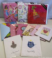 Photo showing eleven birthday cards arranged fanned out. Some cards are in original sealed plastic packaging, others are open with visible envelopes. Designs include dancing women, whimsical animals, party scenes, hearts and humorous illustrations.