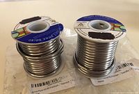 Two rolls of silver-colored stained glass solder wire on spools with blue and white labels showing product details and stained glass illustration.