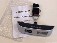 Digital luggage scale with LCD display, unit button, power button, strap, metal hook, and user manual on bubble wrap.
