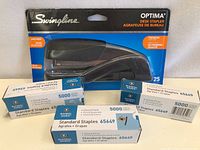 Photo showing one black Swingline Optima desk stapler in packaging along with five boxes of standard staples.