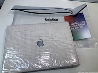 Top front view of textured basket weave MacBook cover with Apple logo visible, inside Uniqfind clear zip bag with brand name and card.