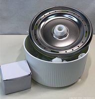 View of water pet fountain showing stainless steel top with water outlets and white plastic base, with small white box beside it.