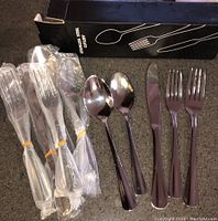 Photo showing some cutlery pieces, including knives, forks, and spoons with some wrapped in plastic and a black box labeled stainless steel cutlery in the background.