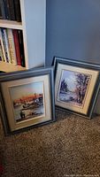 Two original framed paintings by the same artist leaning against a wall, showing their general size and matching frame style.