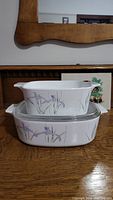 Two stacked white Corning Ware Shadow Iris casserole dishes, larger one with glass lid, showing purple iris flower pattern on side.