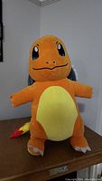 Front view of large Charmander plush toy with bright orange body and yellow belly, standing on a wooden surface.