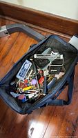 Top-down view inside the open tool bag showing multiple hand tools and accessories inside the bag on a wooden floor.