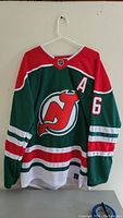 Front view of green New Jersey Devils hockey jersey with red shoulders, white stripes on sleeves and waist, large Devils logo on the chest, and 'A' alternate captain patch on the upper left chest.