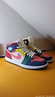 Front-side view of the Nike Air Jordan 1 Mid sneakers showing pink, green, yellow, navy blue, red, and white panels with white laces.