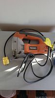 Side angle of Ridgid jigsaw showing orange body, black handle, and power cord with yellow plug.