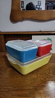 Blue, red, and yellow rectangular Pyrex refrigerator dishes stacked with clear glass lids on a wooden surface.
