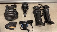 Photo showing baseball catcher's gear including chest protector, helmet, leg guards, glove, and protective cup arranged on floor.