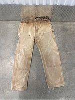 Front view of heavy-duty beige Carhartt work pants, showing wear and dirt stains, reinforced knees, and utility pockets including hammer loop.