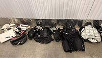 Photo of the overall spread of hockey goalie equipment on the floor against a metal wall, including pads, gloves, chest protector, and pants.