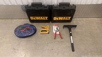 Two black Dewalt tool cases, blue air hose in plastic bag, yellow staple gun, pliers, punch tool in packaging, ice scraper with brush, all displayed on floor.