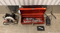 Wide shot showing electric circular saw, red metal toolbox with hand tools, black charger, and a small black blade case on cement floor.