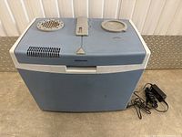 Blue and grey Mobicool electric cooler shown on concrete floor with power adapter next to it.