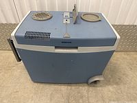 Image of a blue Mobicool Cooler S portable cooler with a white lid containing ventilation holes, control panel, and two round panels. One wheel is visibly broken on one side of the cooler. The cooler is placed on a concrete floor.