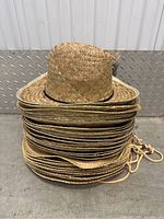 Stack of approximately twenty woven straw cowboy hats with natural tan color, black bands around crowns, and chin straps, against a metal diamond plate background.