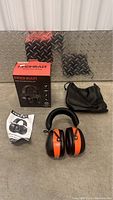 ProHear hearing protector headphones with orange and black ear cups, padded headband. Includes original packaging box, black carrying pouch, and user manual laid out on diamond plate metal floor.