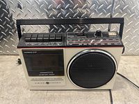 Front view showing radio speaker, cassette compartment, multiple control buttons on top, tuning dial with frequency markings, and carry handle.