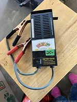 Top-down view of battery tester on wooden surface showing the tester face with the analog meter, cables with red and black clamps, and the toggle switch.