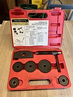 Open brake caliper tool kit showing T-handle tool and five adapter discs plus one rectangular plate inside red molded plastic case.
