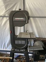 Full view of the Dremel band saw on its stand inside a tent, showing black metal frame and work table.