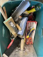 Photo of a green plastic tote filled with various drywall tools including multiple drywall knives, sanding block, corner tool and drywall tape.