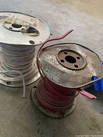 Photo showing two metal spools containing thick insulated electrical wire in red and white colors on a concrete floor.