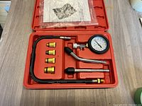 Open red plastic case displaying engine compression tester components including dial gauge, hoses, adapters, and hand pump.