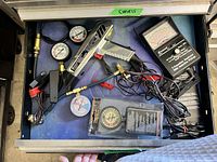 View of a drawer containing two testers: a Simpson 260-8 analog multimeter and a digital temperature tester, along with multiple pressure gauges, a handheld tool with nozzle, temperature probe, and assorted cables and connectors. The items are arranged loosely in the drawer.