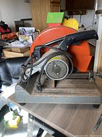 Front and left side view of the Makita cut-off saw showing handle, motor, large orange blade guard, mounted on metal base with attached cord.