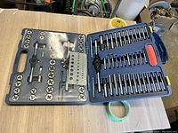 Open blue plastic molded case with assorted taps, dies, tap holders, and threading guide block visible, placed on workbench surface.
