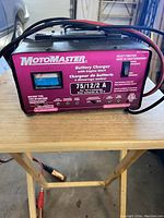 Front view of Motomaster battery charger on wooden table, showing model number, amp meter, function selector switch, and cables attached.