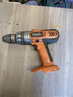 Photo showing a single Rigid cordless drill lying on a wooden surface, showing orange and black body with adjustable torque collar and worn metal chuck.