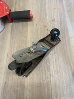 Top and side view of Stanley hand planer showing black front and rear knobs, metal body, adjustment lever, and blade plate with branding.