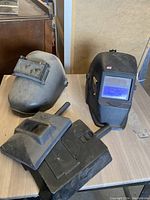 Photo showing black auto-darkening welding helmet, gray fixed-shade welding mask, and black hand-held welding face shields on a wooden surface.