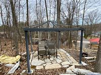 Metal gazebo frame without canopy set on a stone paved cleared area in a wooded outdoor environment. Four stacked outdoor chairs under the frame.