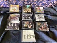 Overview of 10 Beatles CDs laid out on dark fabric background with blue patterned fabric backdrop. Shows album cover art for each CD case in jewel case packaging.