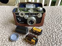 Front view of Argus C3 camera in brown leather case with two rolls of unused film and a film canister on a fabric surface
