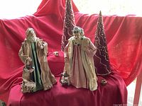 Two vintage Silvestri Santas dressed in fabric robes, two tall red crystal beaded Christmas trees with gold swirl patterns, three small jungle bells on a red fabric background.
