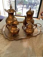 Full 5 piece brass serving set arranged on a patterned tablecloth in front of glass doors, showing water carafe, tea or coffee pot, sugar bowl, creamer, all on tray.