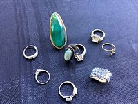Photo showing all nine vintage ladies rings arranged on a dark fabric background, highlighting the large green stone ring and others with clear and blue stones.